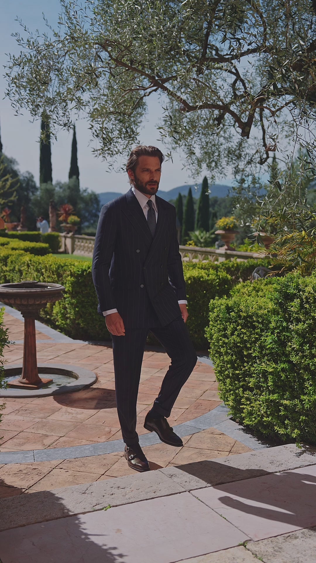 Costum masculin închis la culoare, purtat pe o alee de grădină