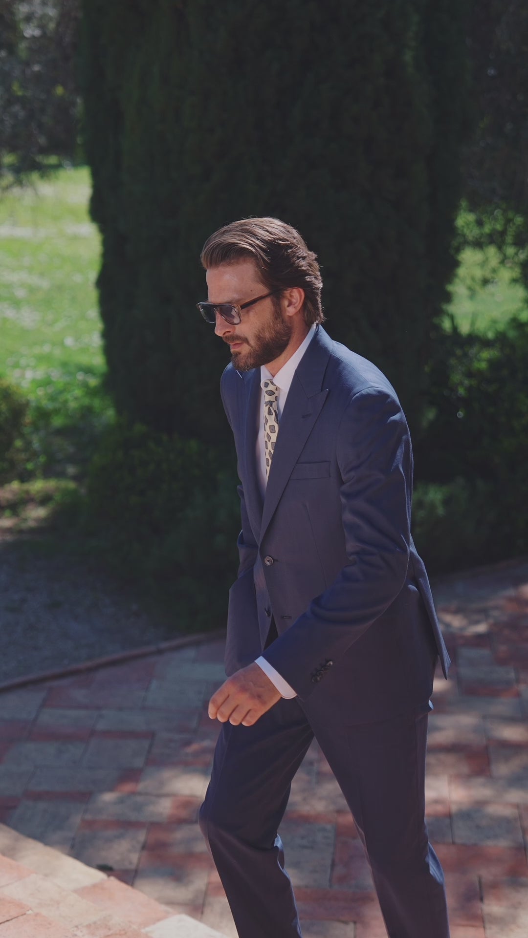 Costum masculin bleumarin cu cămașă albă și cravată, pe o alee umbrită
