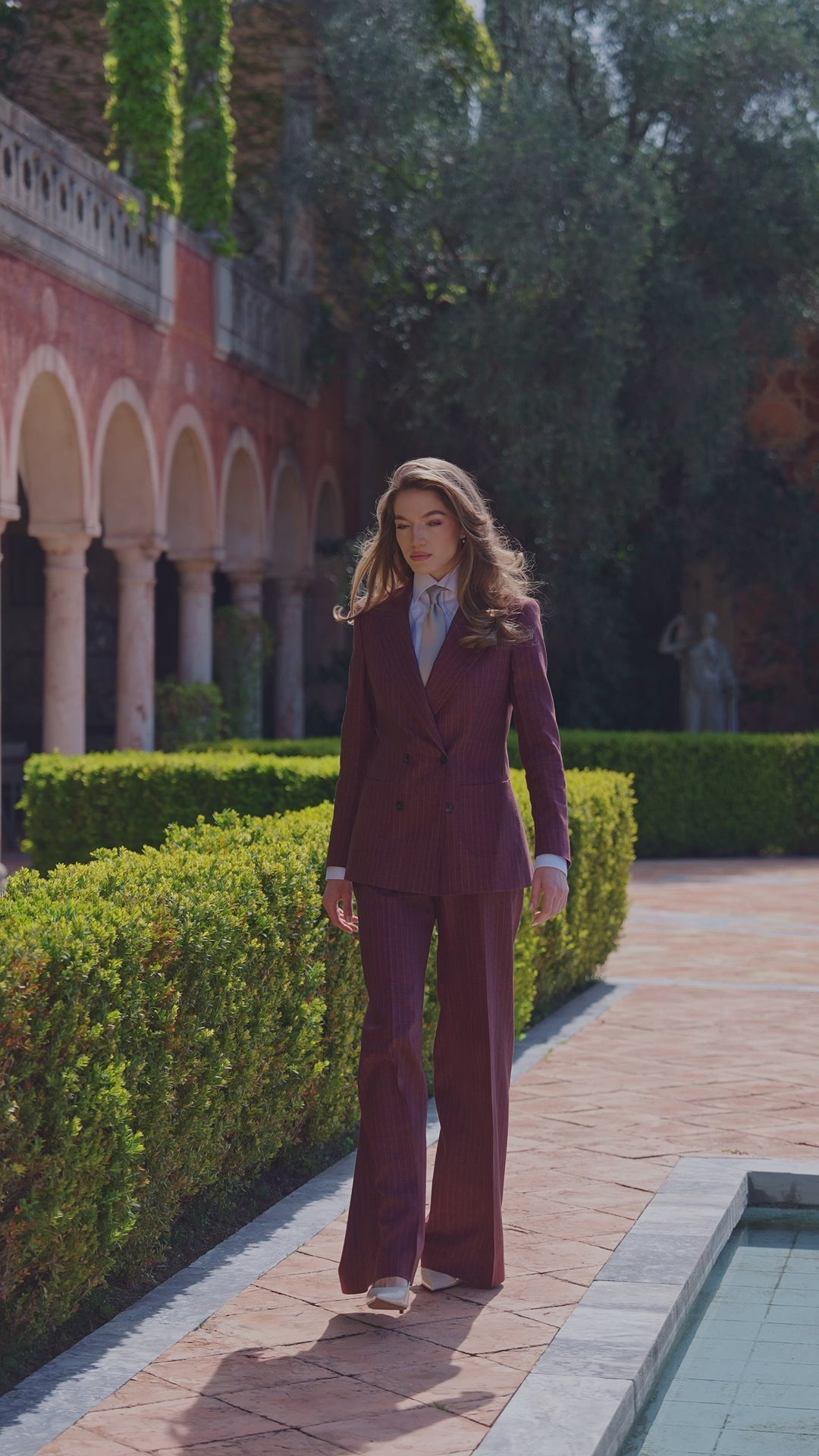 Costum feminin burgund cu pantaloni cu croială amplă, pe o alee de grădină