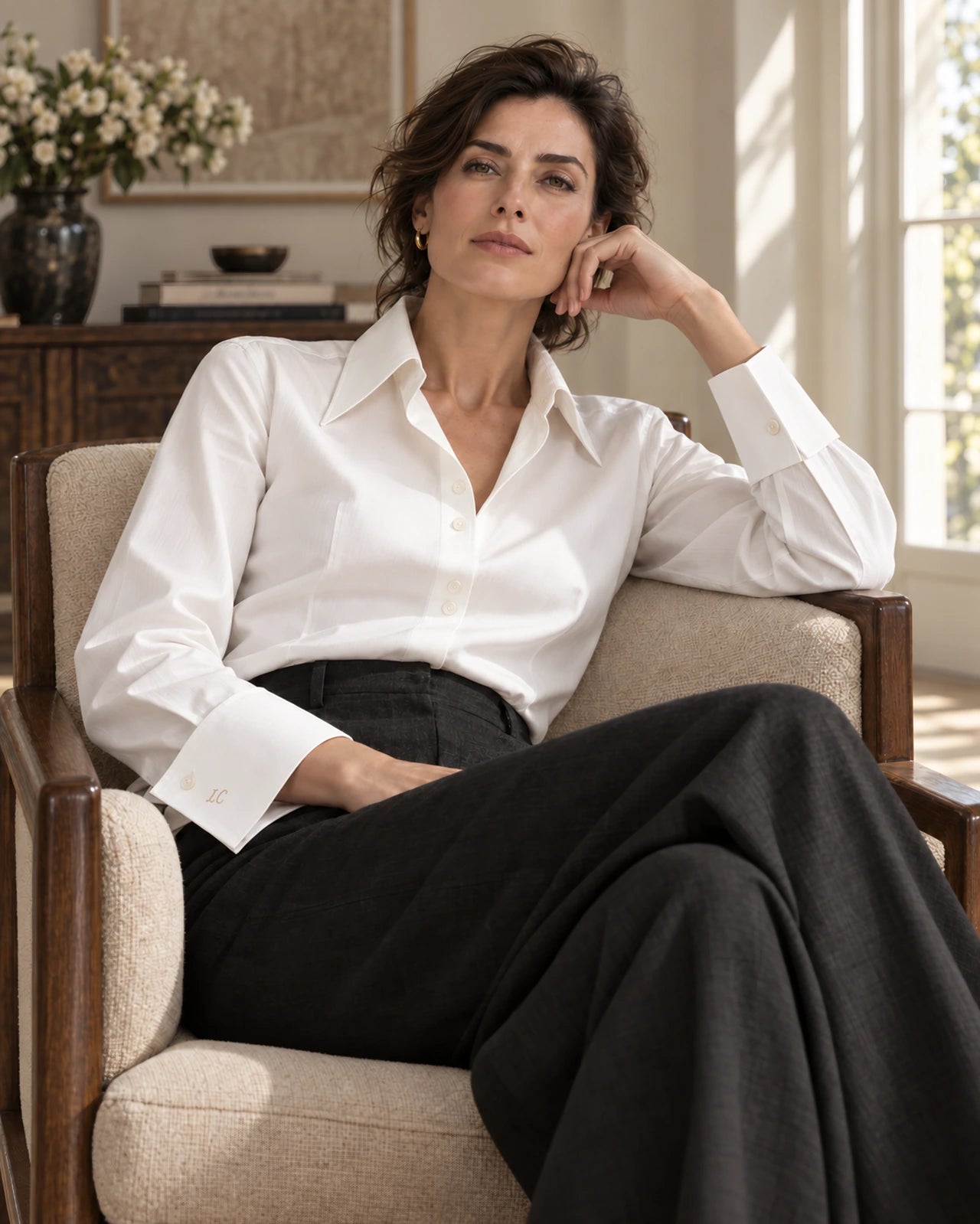 Femeie în cămașă ivoire cu guler amplu și manșete modelate, relaxată într-un fotoliu, cu privirea în cameră și pantaloni antracit.